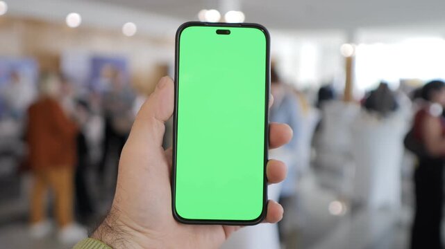 Hand holds smartphone with green screen. Background shows blurred indoor crowd. Suitable for app demos or digital content. Focus on device, not people. Ideal for UI/UX design or marketing visuals