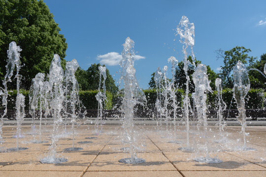 Active water fountain jets spray and splash from a stone pavement in a public city park. A clear blue sky and green trees create a refreshing urban background on a hot summer day.