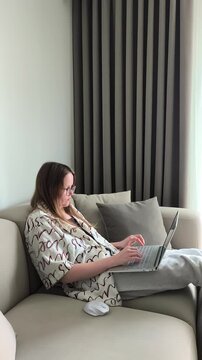 Woman continues typing on her laptop while seated comfortably on the couch at home. She works diligently on her computer during a remote session. Concept of home office, typing and freelance