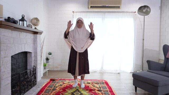 A senior Muslim woman, dressed in traditional Middle Eastern attire, is performing a prayer ceremony to Allah in the prayer room.