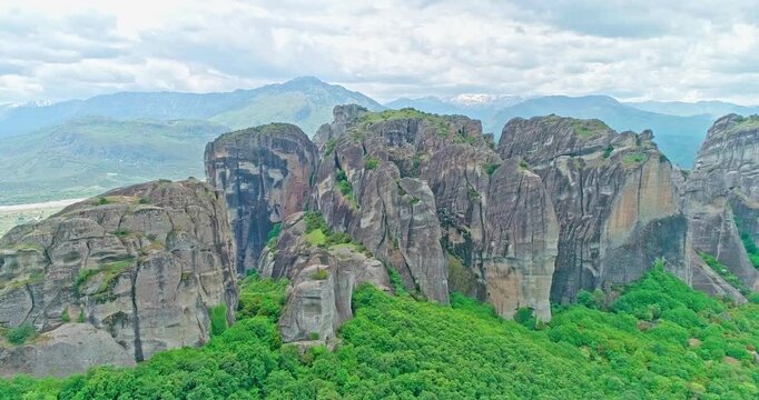 Slow aerial drone push-in toward the Meteora rock formations in Greece, with green forest, cliffs, mountains, and a cloudy sky. Scenic travel footage of the famous UNESCO World Heritage lands