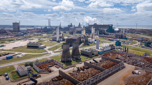 Aerial view of the Tata Steel industrial complex with large smokestacks, storage piles, and railway tracks under a cloudy sky in Velsen-Noord, Noord-Holland, Netherlands.