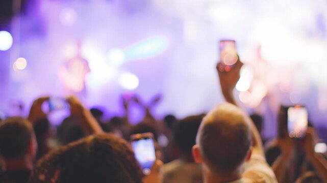 Dense concert audience viewed from behind with blurred silhouettes, and raised hands creating energetic, shared music moment that communicates excitement, anticipation. 