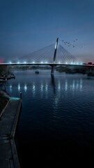 Fototapeta premium Modern cable-stayed bridge over river at dusk with city skyline and boats