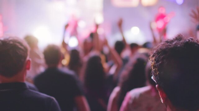 Dense concert audience viewed from behind with blurred silhouettes, and raised hands creating energetic, shared music moment that communicates excitement, anticipation. 
