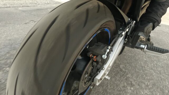 Low rear side view of motorcycle wheel brake assembly with rider boot on footpeg showing rotating tire blur and blue rim accent on paved road with sport performance detail