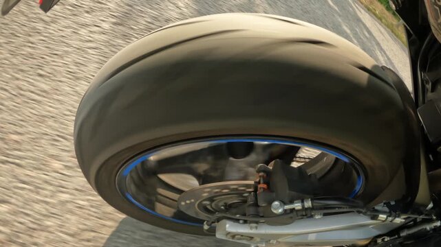 Close low oblique view of rear motorcycle wheel with blue rim stripe, exposed brake caliper, disc rotor, metallic reflections, and detailed motion impression on textured asphalt