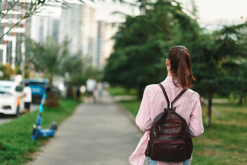 Naklejka premium Woman lifestyle, walking away with backpack along a sunlit city path, casual stride, greenery beside the route, candid moment, relaxed mood, natural colors