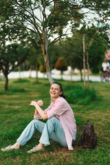 Naklejka premium A smiling young woman student sits on the grass in a park, wearing casual clothes with a backpack nearby. Concept of student life and outdoor leisure.