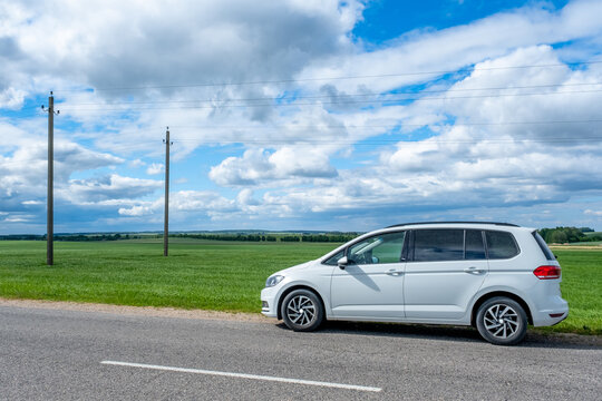 A white minivan is parked on the side of a paved road next to a green agricultural field under a cloudy blue sky