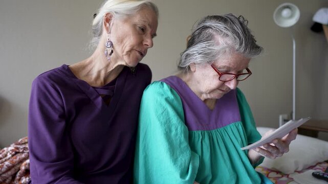 Mature daughter visits her elderly mother at a senior care facility to share stories and look at photos together during their time together.