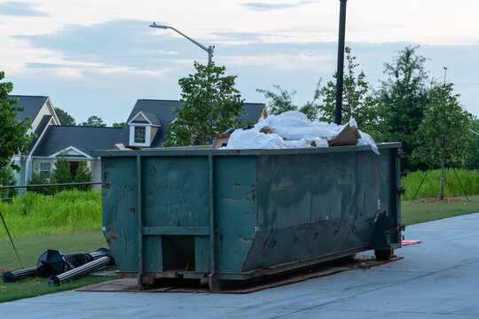 Large green rusty roll-off dumpster overflowing with household trash and construction debris on a residential driveway, indicating a major cleanout or renovation project in a suburban neighborhood.