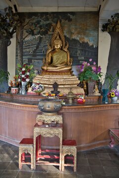 Golden Buddha statue sitting in meditation at Brahmavihara-Arama monastery in Bali, decorated with colorful flowers and a smaller stone statue in front against a painted temple background.