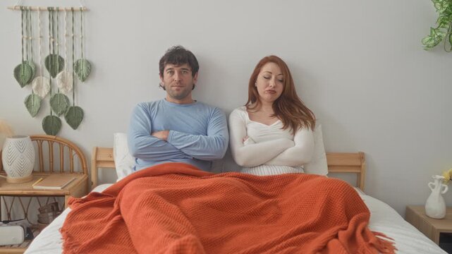 Man and woman sit upright on bed with arms crossed, orange blanket pulled over laps, bedside plants and macrame on wall in bedroom; relationship tension.