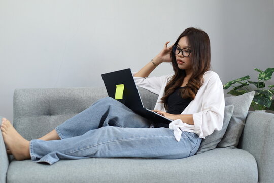 A young Asian woman sits on the sofa in her living room, working on her laptop while holding her head with a tired, lethargic and stressed expression due to a work deadline.