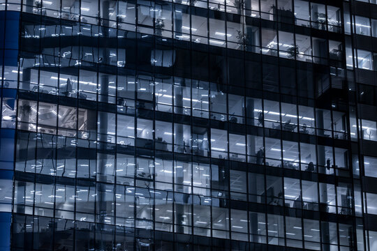 Glass windows across an office building facade at night expose corporate interior floors, while urban architecture curves through cool blue reflections
