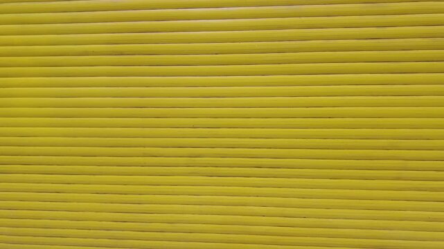 The camera moves backward from a bright yellow metal roller shutter door. The physical movement gradually reveals the structured horizontal texture of the protective security blinds.