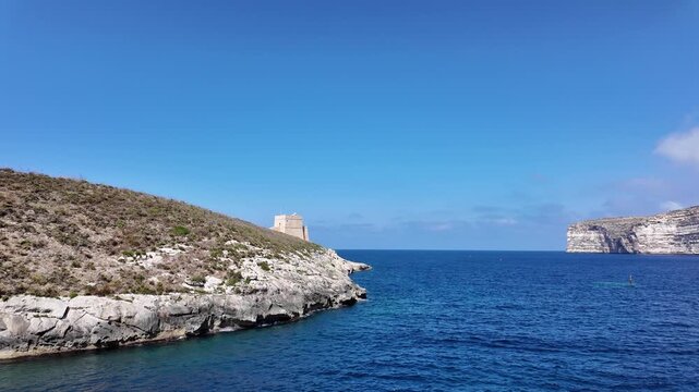 Low angle view of the Xlendi Tower standing against the azure sea and rugged coastline, creating a stunning contrast, Xlendi, Munxar, Malta. High quality 4k footage