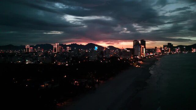 Da nang city coastal panorama featuring bright building lights illuminating the urban skyline and beachfront at dusk, set against mountains as the last hues of sunset fade into the dark sky