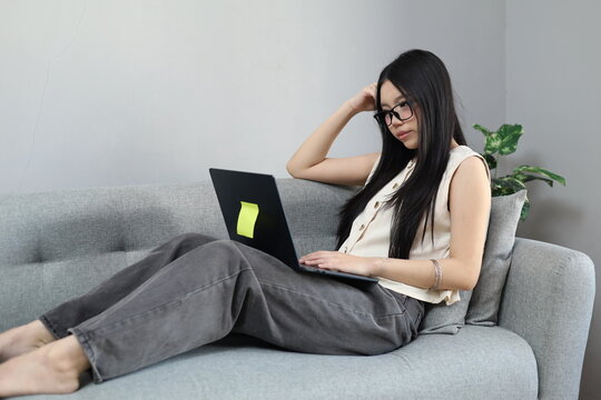 A young Asian woman sits on the sofa in her living room, working on her laptop while holding her head with a tired, lethargic and stressed expression due to a work deadline.