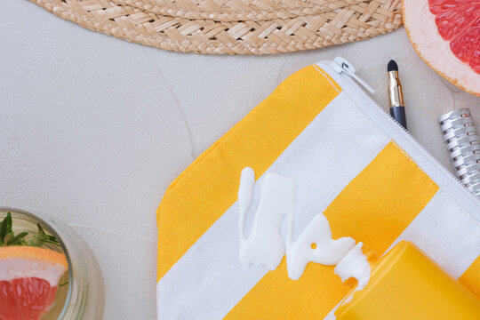 An open cosmetic bag with spilled sunscreen is surrounded by a straw hat, beach sandals, half a grapefruit, a makeup brush, and cosmetic accessories on the sand. top view
