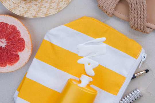 An open cosmetic bag with spilled sunscreen is surrounded by a straw hat, beach sandals, half a grapefruit, a makeup brush, and cosmetic accessories on the sand. top view