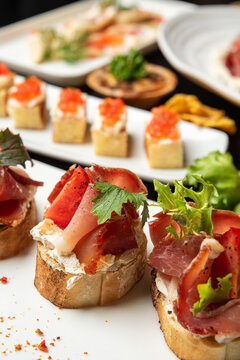 Gourmet bruschetta with prosciutto ham, cream cheese, and fresh greens on white background. Catering appetizers with salmon caviar canapes blurred in the background.