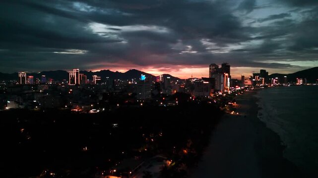 Danang city skyline illuminating at twilight, urban architecture framing a vibrant coastal beach and tranquil ocean waves rolling in, soft sunset glow behind distant mountains