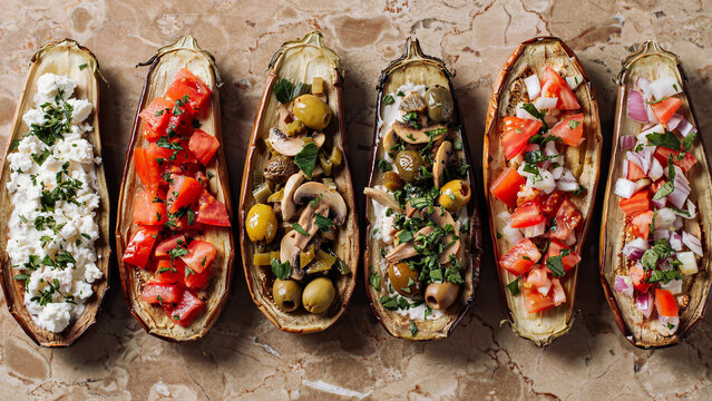 A top-down photo of six roasted eggplant halves arranged in a row on a brown marble surface, each filled with different Mediterranean toppings