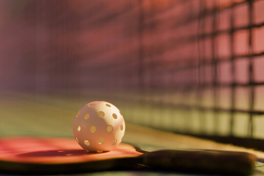 White pickleball with holes resting on a pink paddle on the court, close up with a tennis net blurred in the warm sunset background. Perfect for sports concepts.