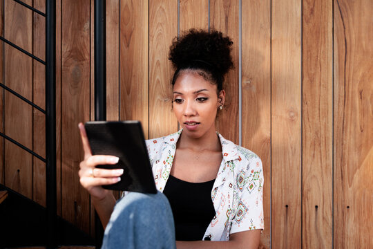 Woman with digital tablet sits by wooden wall and stairs.