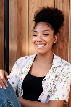 Smiling woman with curly hair sits against wood wall.