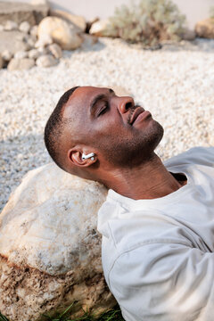 Man with earbuds rests head on rock outdoors.