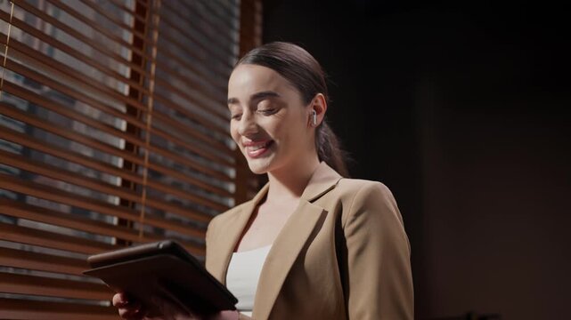 Close up of woman standing near window blinds. Female in beige jacket and pony tail. Movie agency worker using wireless headphones. Smiling female casting director holding tablet.