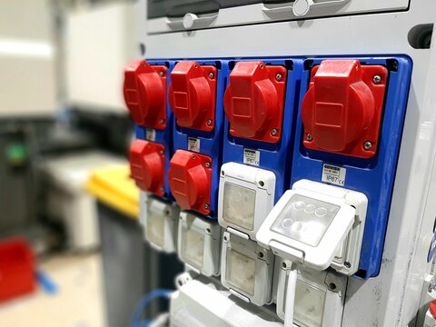 Industrial three phase power plugs and sockets on factory wall. Electrical cables connected to heavy duty red high voltage outlets in workshop background.