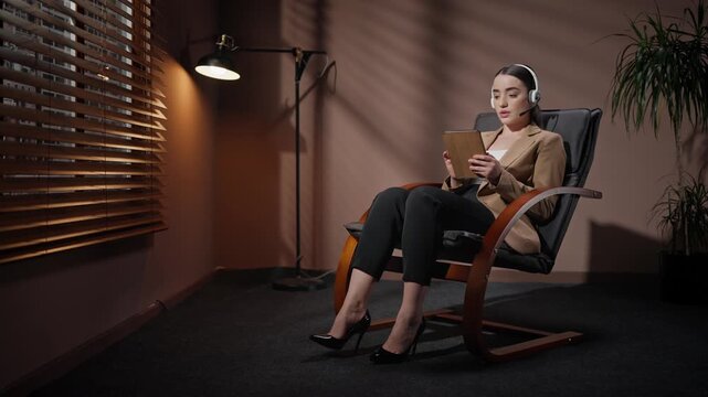 Female conducting video conference. Woman sitting on chair and conducting video chat. Female wearing headphones and talking in microphone. Woman holding tablet in dim loft room.