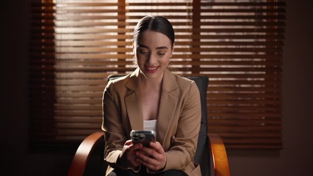 Front view of woman sitting at background of window blinds. Female smiling and looking at smartphone screen. Woman chatting isolated inside dark room. Happy woman using social media.
