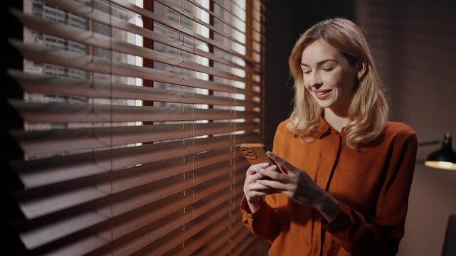 Female casting director wearing orange shirt. Woman isolated near window blinds. Happy woman smiling standing in dark room. Blonded woman holding smartphone and texting message.
