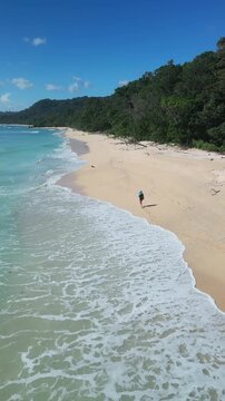 Asian woman hiking along remote beach drone perspective with mountain backdrop lush forest edge long curve of sand rhythmic waves footprints leading into distance adventurous, Java island