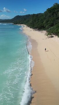 Aerial turquoise beach woman walking along shoreline under jungle cliffs, solitary traveler leaving footprints in soft sand as gentle waves lap coast, driftwood and rocky outcrops, vertical video