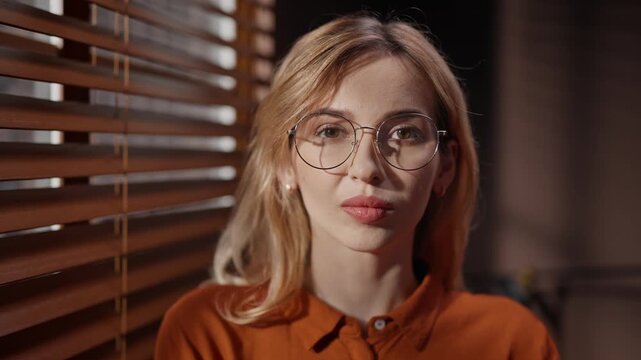 Portrait of woman attentively looking through window blinds. Female wearing glasses and orange shirt. Woman turning head and looking at camera. Casting director at audition room.