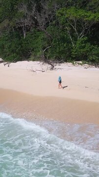 Aerial turquoise shore woman with backpack walking near driftwood and gentle surf, wide crescent of pale sand meets clear ocean beneath lush hills, relaxed vacation pace, vertical video