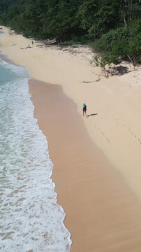 Aerial view woman exploring tropical shoreline, emerald surf rolling onto pale sand, dense jungle backdrop, footprints leading into distance, bright sky, freedom and travel discovery