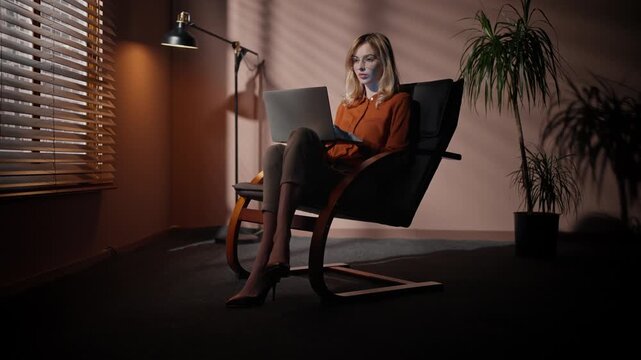 Professional woman crossing legs on office chair. Female sitting at chair at background of dim design. Woman in official suit sitting at dark room. Female director using laptop in audition room.