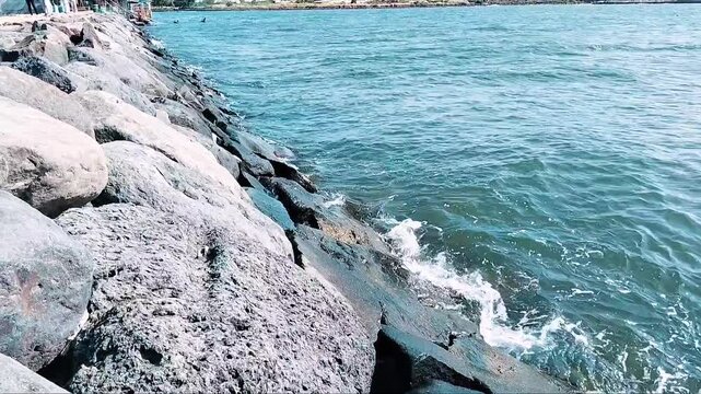 Ocean waves crashing against a concrete breakwater under a clear blue sky. The coastal protection structure stretches into the sea, creating a peaceful yet dynamic seascape with beautiful ocean views.
