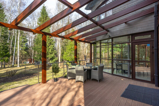 The scene shows an outdoor deck area with a seating arrangement made of wicker. Sunlight shines on the deck while trees surround the backyard. A house is nearby, adding to the setting