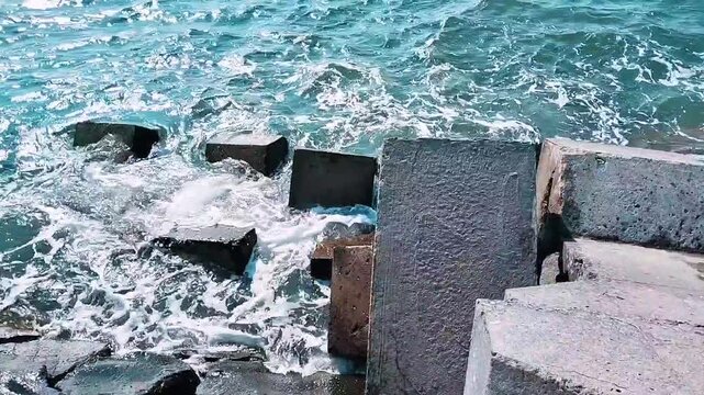 Ocean waves crashing against a concrete breakwater under a clear blue sky. The coastal protection structure stretches into the sea, creating a peaceful yet dynamic seascape with beautiful ocean views.