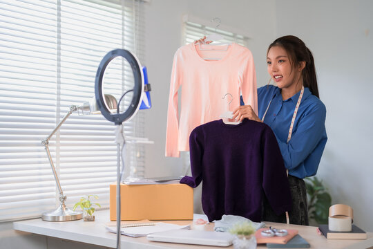 Young Asian woman online merchant presenting clothing items to camera while streaming fashion sale in home office