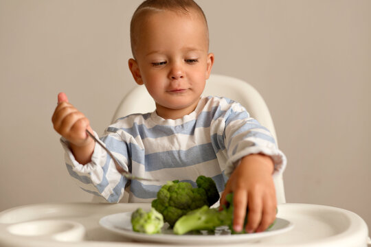 A young child sits at a table, using a spoon to eat broccoli from a plate. The child appears focused on the food, enjoying the mealtime
