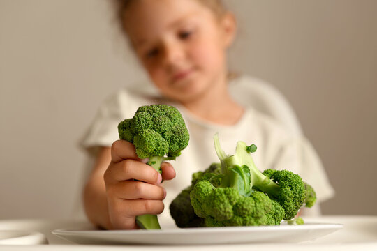 A child is examining a piece of broccoli while sitting at a table with a plate of green vegetables. It is an afternoon meal time at home